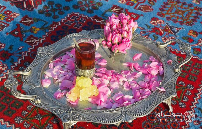 یک لیوان چای، چند نبات و تعدادی غنچه و گلبرگ گل محمدی در سینی نقره‌ روی قالی آبی‌رنگی قرار داده شده‌اند.